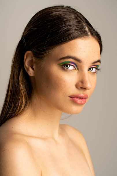 Подростковая модель с обнаженными плечами и красочными тенями для век, смотрящая на камеру, изолированную на сером  - Фото, изображение