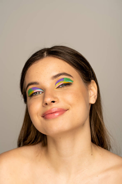 Радостная модель-подросток с разноцветным макияжем, смотрящая на камеру, изолированную на сером  - Фото, изображение