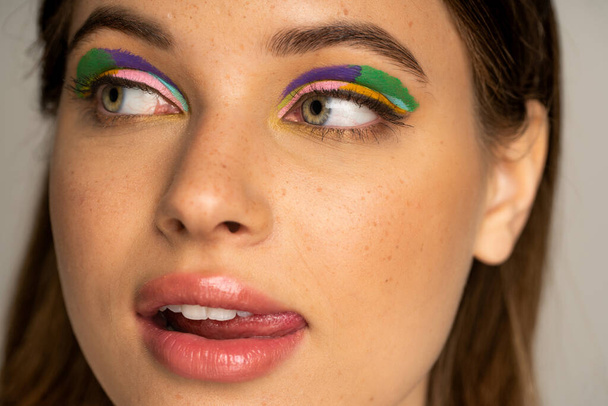 Close up view of teen model with multicolored visage sticking out tongue isolated on grey  - Foto, afbeelding