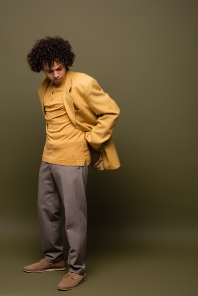 full length of african american man in yellow blazer and trousers posing with hand behind back on grey green background - Valokuva, kuva