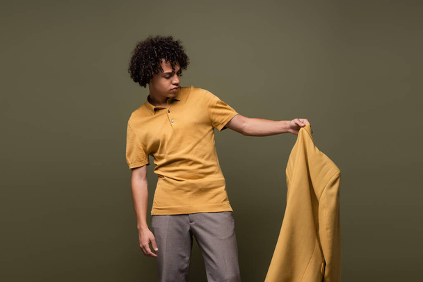 κομψό αφροαμερικανός άνδρας σε πόλο πουκάμισο και παντελόνι κρατώντας κίτρινο σακάκι απομονώνονται σε γκρι - Φωτογραφία, εικόνα