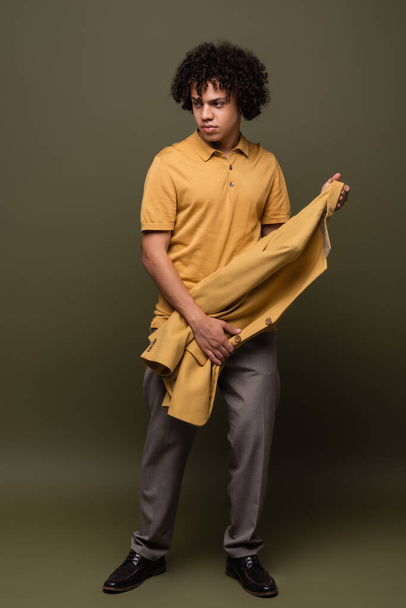 full length of young and trendy african american man in yellow polo shirt holding blazer on olive grey background - Foto, immagini