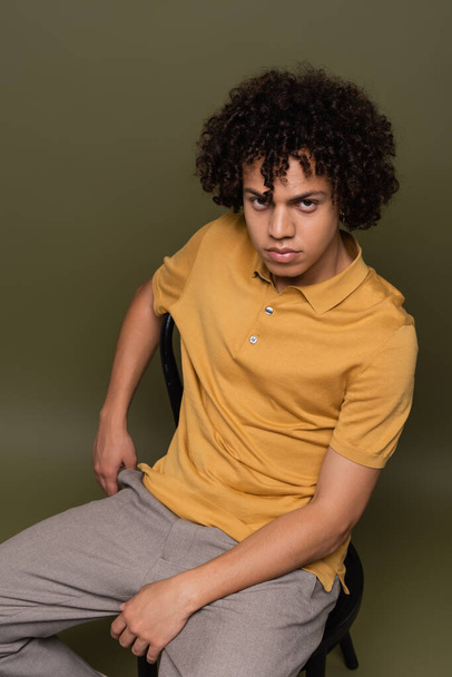 jong Afrikaans amerikaans man in gele polo shirt zitten op stoel en kijken naar camera op grijs groene achtergrond - Foto, afbeelding