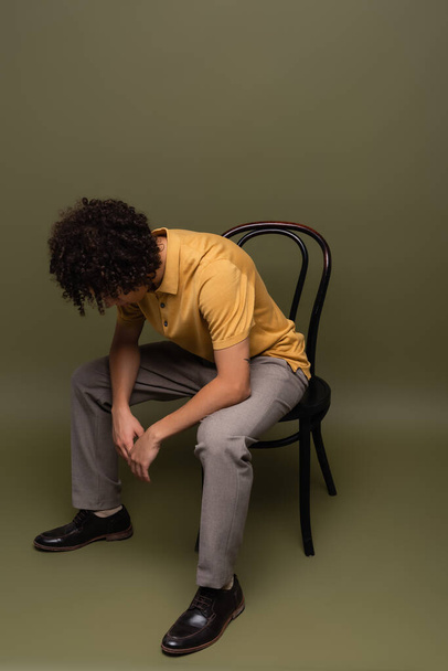 full length of african american man in yellow polo shirt and black leather shoes sitting on chair with bowed head on grey green background - Foto, immagini