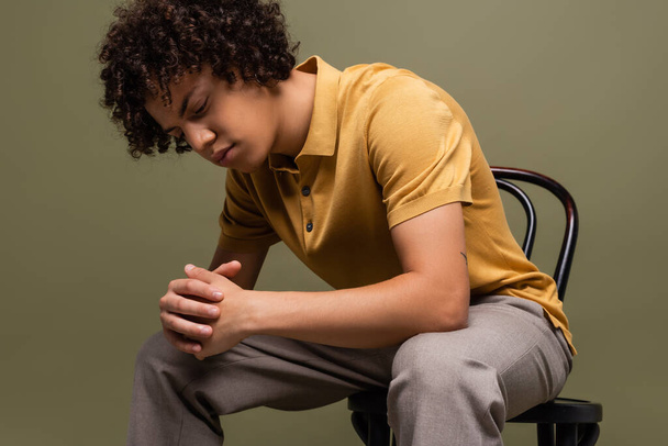young african american man with curly hair wearing yellow polo shirt and sitting on chair with clenched hands isolated on grey - Photo, Image