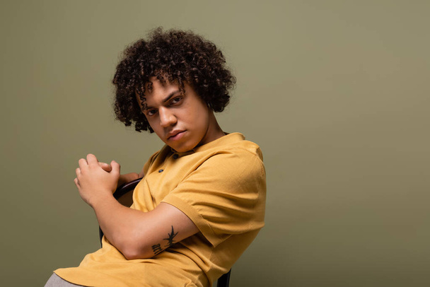young tattooed african american man in yellow polo shirt looking at camera while posing on chair isolated on grey - Photo, image