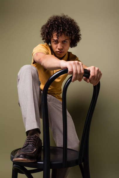 young african american man in trendy casual attire stepping on chair and looking at camera on grey green background - Foto, immagini