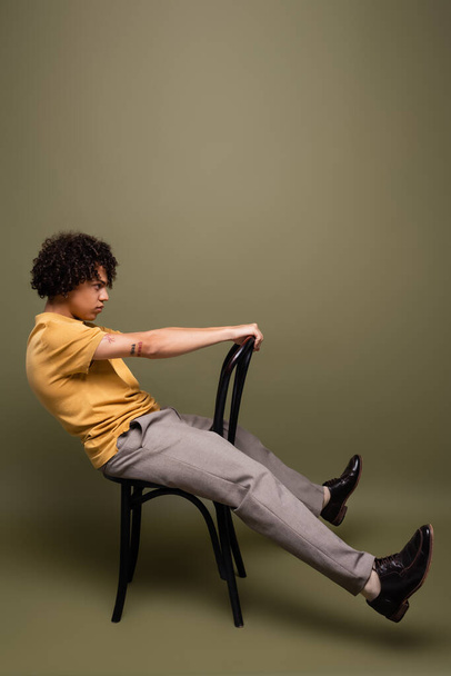 vue latérale de l'homme afro-américain tatoué en t-shirt jaune et chaussures en cuir noir posant sur chaise sur fond gris-vert - Photo, image