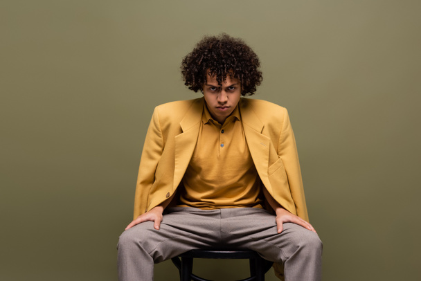 front view of fashionable african american man in yellow blazer and trousers sitting and looking at camera on grey background - Foto, immagini
