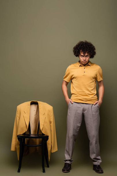 longitud completa del joven afroamericano de pie con las manos en los bolsillos cerca de la silla con chaqueta amarilla sobre fondo verde gris - Foto, imagen