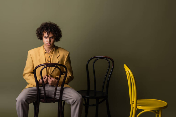 african american guy in stylish outfit posing with black and yellow chairs on olive grey background - 写真・画像