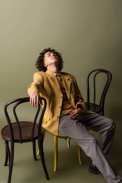 joven y moderno hombre afroamericano con los ojos cerrados sentado cerca de sillas negras sobre fondo gris oliva - Foto, imagen
