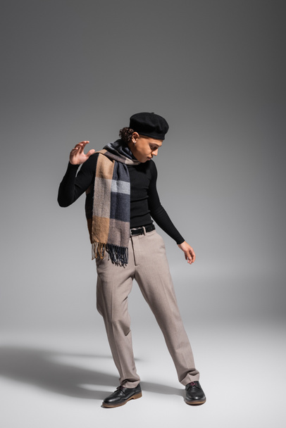 full length of stylish african american man in black sweater and plaid scarf with beret posing on grey background - Photo, Image