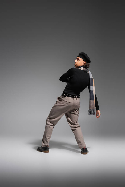 pleine longueur de l'homme afro-américain en béret noir et col roulé avec écharpe à carreaux regardant loin sur fond gris - Photo, image