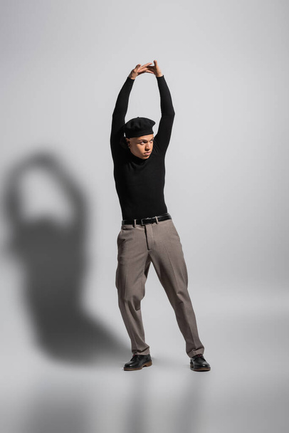 full length of african american man in black turtleneck and trousers with leather shoes standing with raised hands on grey background - Photo, image