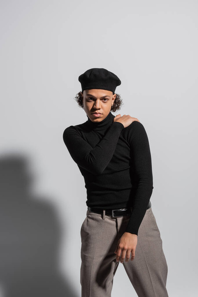 african american man in fashionable beret and black turtleneck posing with hand on shoulder on grey background - Photo, Image
