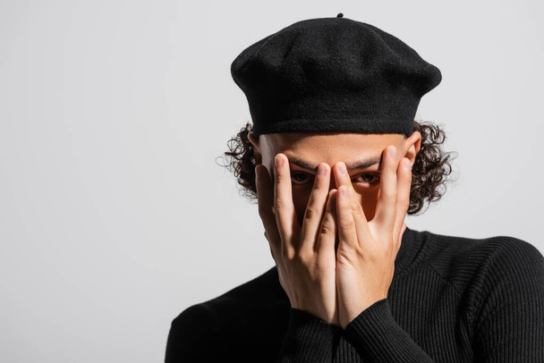young african american man in black stylish beret obscuring face with hands and looking at camera isolated on grey - Photo, image
