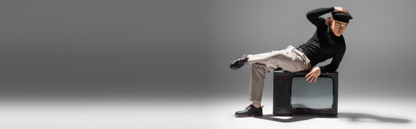 comprimento total do homem americano africano em traje casual elegante tocando boina preta enquanto sentado na tv vintage set em fundo cinza, banner - Foto, Imagem