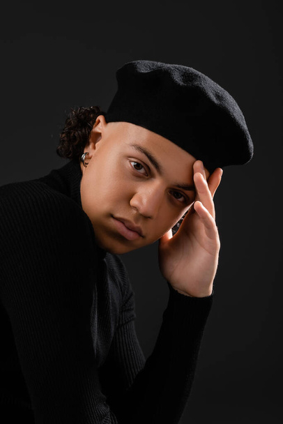 portrait of stylish african american guy in beret and turtleneck holding hand near face and looking at camera isolated on black - Foto, immagini