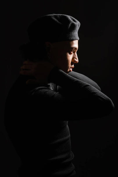 πλαϊνή άποψη του Αφροαμερικανού άνδρα σε πουλόβερ και μπερέ ποζάρουν με τα χέρια πίσω από το λαιμό απομονώνονται σε μαύρο - Φωτογραφία, εικόνα