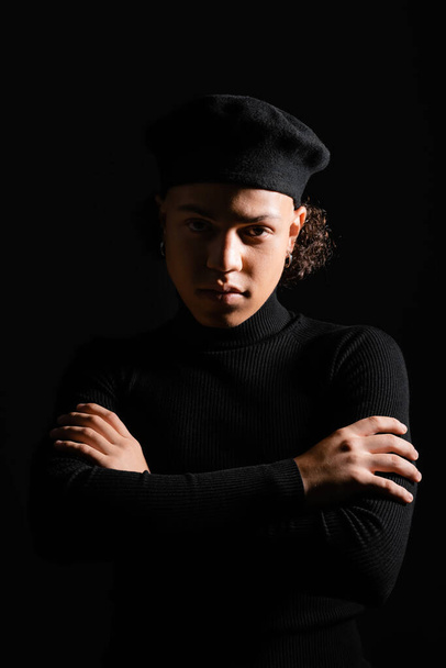 hombre afroamericano con estilo en boina y cuello alto de pie con los brazos cruzados y mirando a la cámara aislada en negro - Foto, imagen