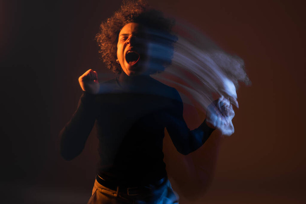 bewegingsvervaging van Afro-Amerikaanse man met bipolaire stoornis en gewond gezicht schreeuwen met gesloten ogen op donkere achtergrond met oranje en blauw licht - Foto, afbeelding