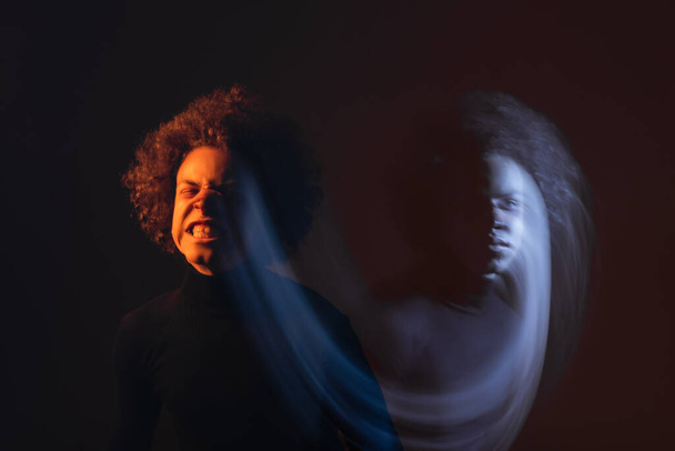 doble exposición del hombre afroamericano con trastorno bipolar y cara lesionada haciendo muecas sobre fondo negro con luz naranja y azul - Foto, Imagen