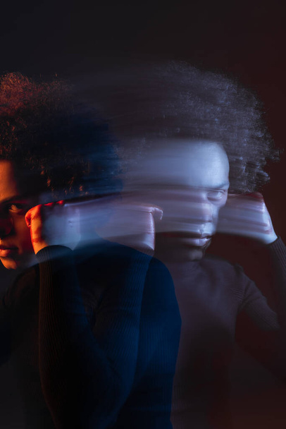 dubbele blootstelling van misbruikt Afrikaans-Amerikaanse man met de hand in de buurt van gewond gezicht kijken naar camera op donker met rood en blauw licht - Foto, afbeelding
