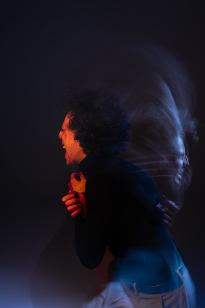 dubbele belichting van gewond en boos Afrikaans amerikaanse man schreeuwen op zwarte achtergrond met rood en blauw licht - Foto, afbeelding