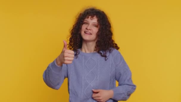 Like. Pretty curly haired woman in sweater raises thumbs up agrees with something or gives positive reply recommends advertisement likes good. Young girl isolated alone on yellow studio background - Séquence, vidéo