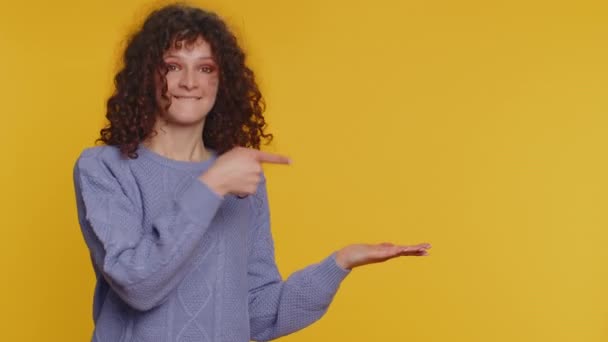 Lovely woman showing thumbs up and pointing empty place, advertising area for commercial text, copy space for goods promotion. Young curly haired girl. Indoor studio shot on yellow wall background - Filmagem, Vídeo