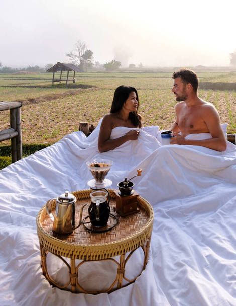 A couple of men and women at a cottage homestay in an outdoor bed in Northern Thailand Nan Province, couple waking up during sunrise in the rice fields in the morning drinking coffee outside - Photo, image