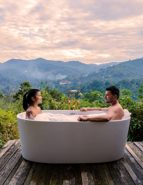 A couple of caucasian men and Asian women in a bathtub in the evening during sunset, a couple in a bathtub. - Foto, Imagen