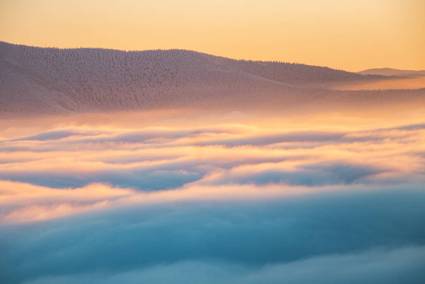 View of sea of clouds colored in the soft orange-pink hue of the morning sun. Peaks of the mountains rising out of this impermeable curtain. Sense of immortality and bliss. Beskydy, Czech republic. - Foto, Imagen