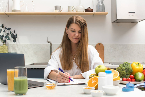 A nutritionist writes an appointment for vitamins and supplements and draws up a nutrition plan during an online consultation with a client, sitting at a table with fresh fruits and vegetables. - Fotoğraf, Görsel