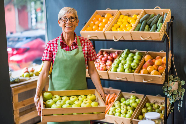 Mature woman works in fruits and vegetables shop. She is holding basket with apples. - Valokuva, kuva