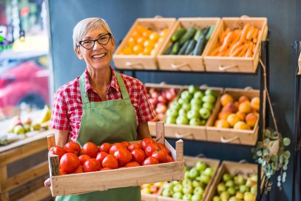 Mature woman works in fruits and vegetables shop. She is holding basket with tomatoes. - Fotoğraf, Görsel