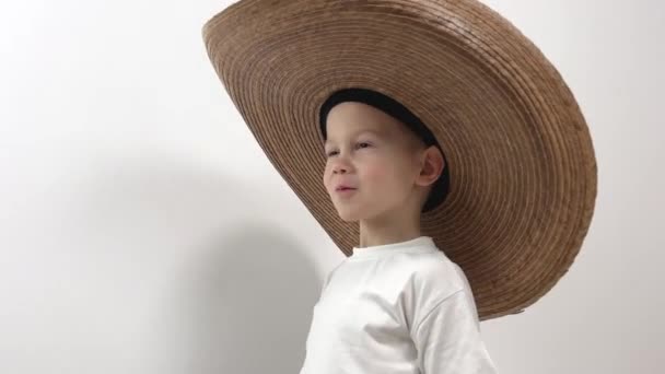  kleine jongen 5 cowboy hoed grote hoed past over de ogen hij rechttrekt het t-shirt is geïsoleerd in studio witte achtergrond hij glimlacht spinnen maakt zijn ogen mooi jongen plaats voor tekst kleding hoofddeksel - Video