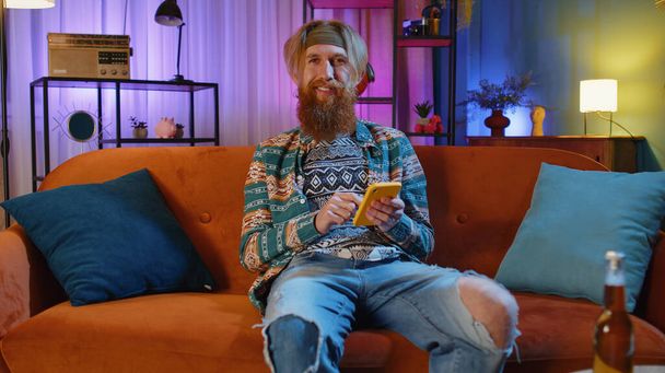 Bearded hippie man sitting on sofa uses mobile phone smiles at night home apartment. Young guy texting share messages content on smartphone social media applications online, watching relax movie - Photo, Image