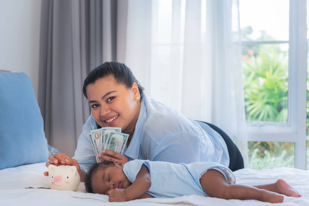 Bambino neonato mezzo thailandese, mezzo nigeriano, 2 mesi dorme su un letto bianco e la madre sta mettendo soldi in un salvadanaio, il concetto è quello di risparmiare soldi per il futuro del bambino.. - Foto, immagini