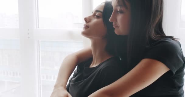 Low angle handheld shot of young lesbian couple smiling and touching hands gently while sitting on sofa against window in light living room at home. Two women sharing love and support holding hands. - Video