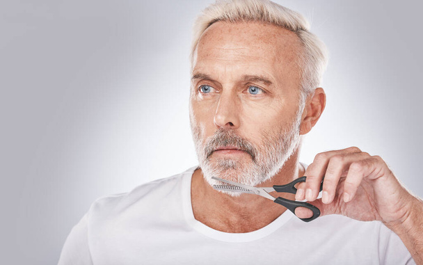 Facial hair, cutting and man with scissors for grooming, shaping and routine treatment on a studio background. Barber, cleaning and face of a senior model trimming beard during morning clean. - Fotoğraf, Görsel