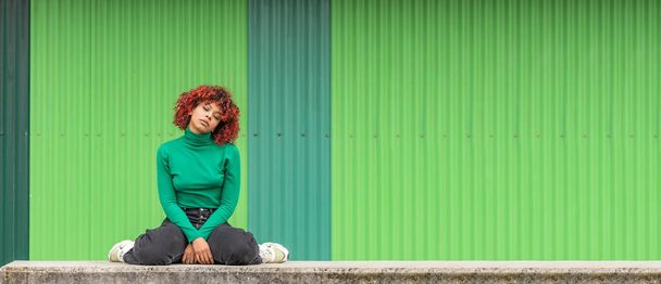 κουρασμένη κοπέλα κάθεται στο δρόμο με πράσινο τοίχο στο παρασκήνιο - Φωτογραφία, εικόνα