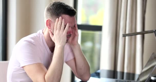 Frustrated freelancer sitting at home in front of laptop and closing eyes. Man overworked by computer feels sudden headache - Video