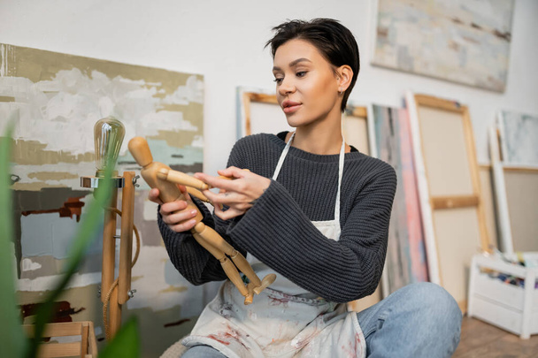 Artist in apron holding wooden doll near blurred paintings in studio  - Foto, imagen