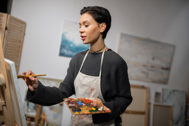 Brunette artist in apron painting on canvas in workshop  - Photo, image