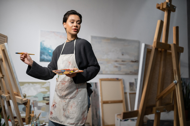 Short haired artist in apron holding paintbrush and palette near canvases on easels in studio  - Photo, image