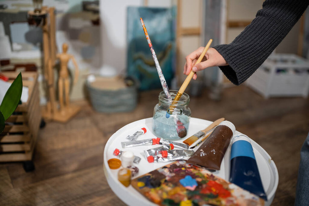 Cropped view of artist taking paintbrush from jar with water near paints in studio  - Foto, Imagen