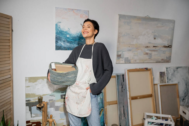 Веселый художник в фартуке держит корзину с оборудованием в мастерской  - Фото, изображение