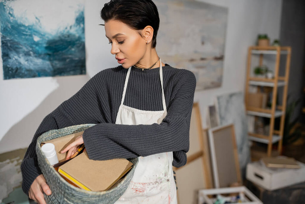 Short haired artist in apron holding basket with equipment in studio  - Foto, imagen